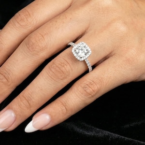 May include: A diamond engagement ring with a square halo setting. The ring features a large, round-cut center stone surrounded by smaller diamonds. The band is also encrusted with diamonds, creating a sparkling effect.