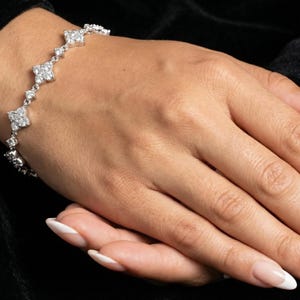 Könnte beinhalten: Ein silbernes Armband mit floralem Design. Das Armband verfügt über mehrere diamantähnliche Steine, die in einem filigranen Muster angeordnet sind. Das Armband wird auf der Hand einer Person vor schwarzem Hintergrund präsentiert.