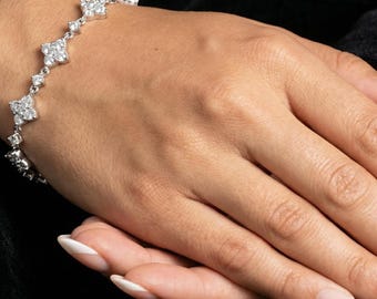 May include: A silver bracelet with a floral design. The bracelet features multiple diamond-like stones set in a delicate pattern. The bracelet is displayed on a person's hand against a black background.