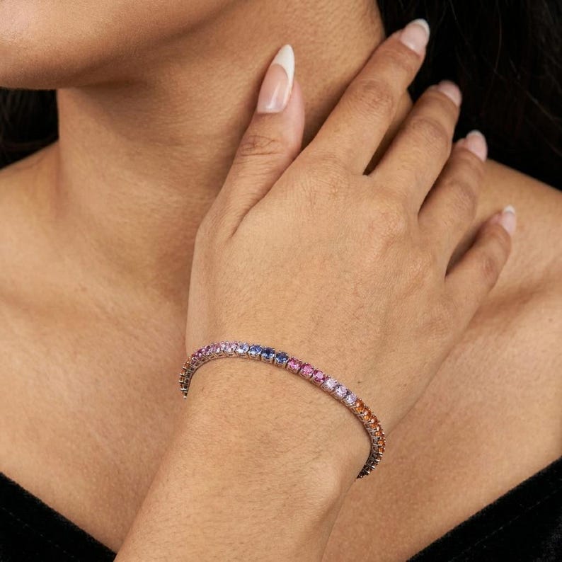 May include: A tennis bracelet featuring a continuous row of round-cut gemstones in a vibrant gradient of colors, transitioning from blue and purple to pink, orange, and yellow, set in a polished metallic band that encircles the wrist.