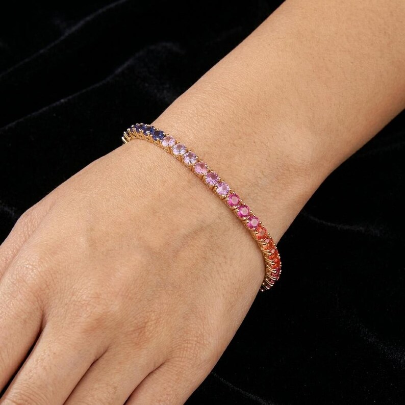 May include: A gold-toned tennis bracelet featuring a continuous row of round-cut gemstones in a vibrant gradient color scheme. The stones transition smoothly from deep blue and purple to pink, orange, and red hues, set in a polished metal frame.