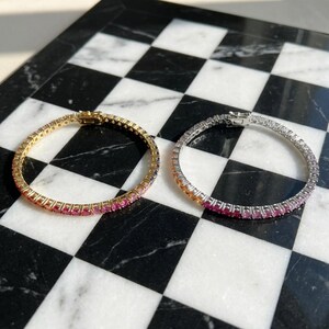 May include: Two tennis bracelets, one in gold-tone metal and one in silver-tone metal, each featuring a continuous row of round-cut gemstones in a gradient of colors transitioning from orange and yellow to pink, purple, and blue hues.