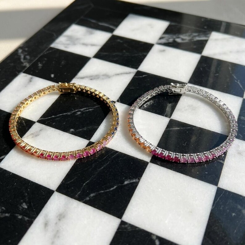 May include: Two tennis bracelets, one in gold-tone metal and one in silver-tone metal, each featuring a continuous row of round-cut gemstones in a gradient of colors transitioning from orange and yellow to pink, purple, and blue hues.