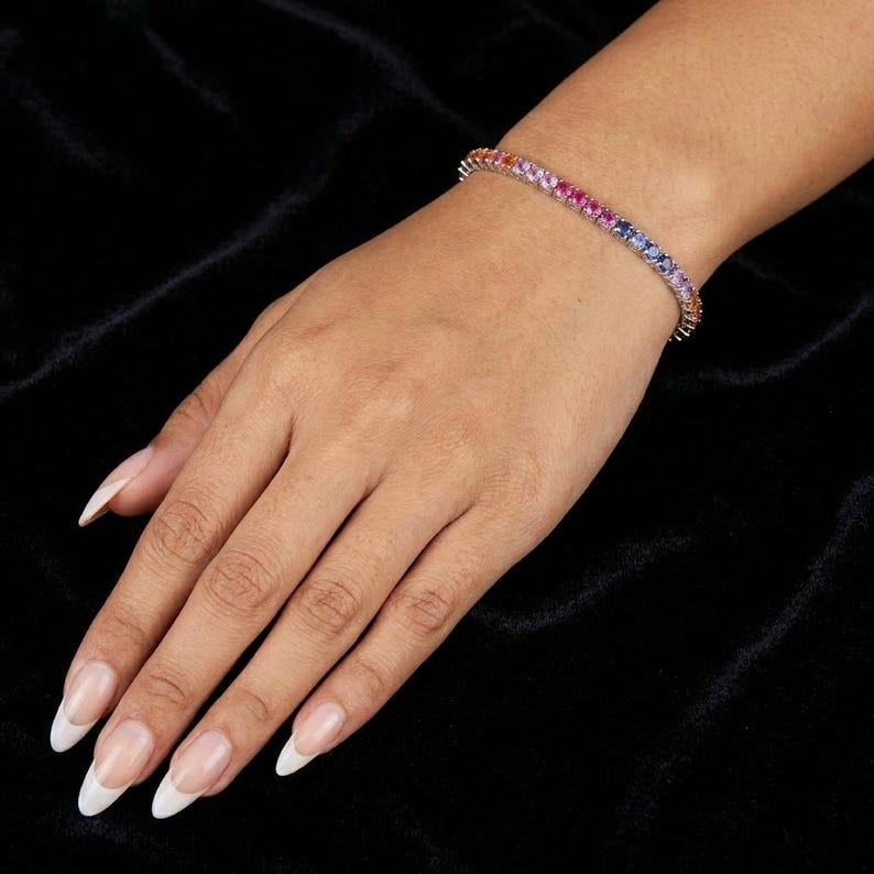 May include: A tennis bracelet featuring a continuous row of round-cut gemstones in a vibrant rainbow gradient, transitioning from orange and pink to purple and blue, set in a polished silver-toned metal band that encircles the wrist.