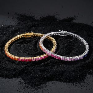 May include: Two tennis bracelets featuring a continuous row of round-cut stones in a vibrant gradient of colors. One bracelet is set in a gold-toned metal, while the other is set in a silver-toned metal, both featuring secure box clasps.