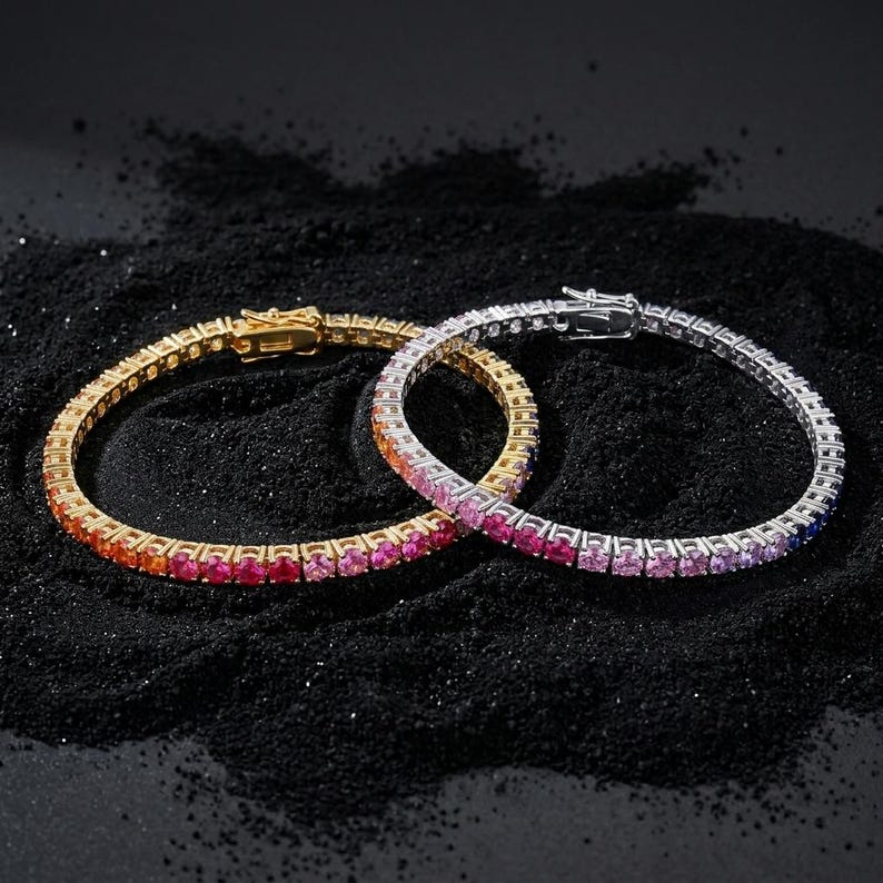 May include: Two tennis bracelets featuring a continuous row of round-cut stones in a vibrant gradient of colors. One bracelet is set in a gold-toned metal, while the other is set in a silver-toned metal, both featuring secure box clasps.