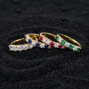 May include: Three gold rings featuring a row of round-cut gemstones. One ring has blue sapphires and clear stones, one has red rubies and clear stones, and one has green emeralds and clear stones. Each ring has a thin, polished gold band.