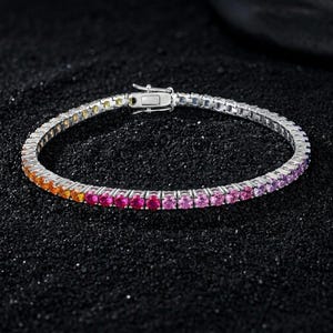 May include: A silver-toned tennis bracelet featuring a continuous row of round-cut stones in a vibrant gradient color scheme, transitioning from yellow and orange to bright pink, magenta, and purple, secured with a rectangular metal box clasp.