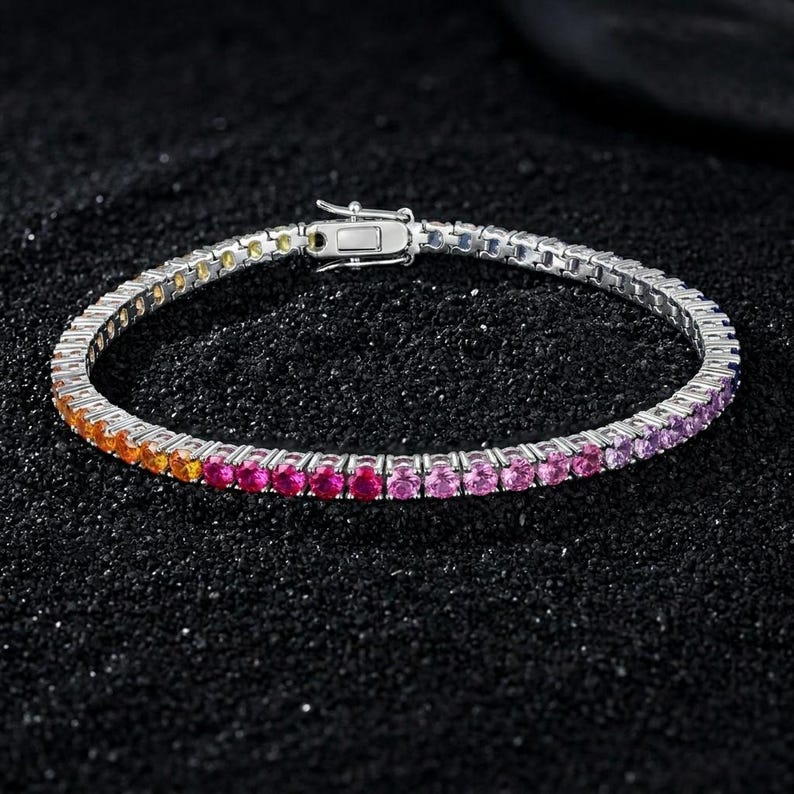 May include: A silver-toned tennis bracelet featuring a continuous row of round-cut stones in a vibrant gradient color scheme, transitioning from yellow and orange to bright pink, magenta, and purple, secured with a rectangular metal box clasp.
