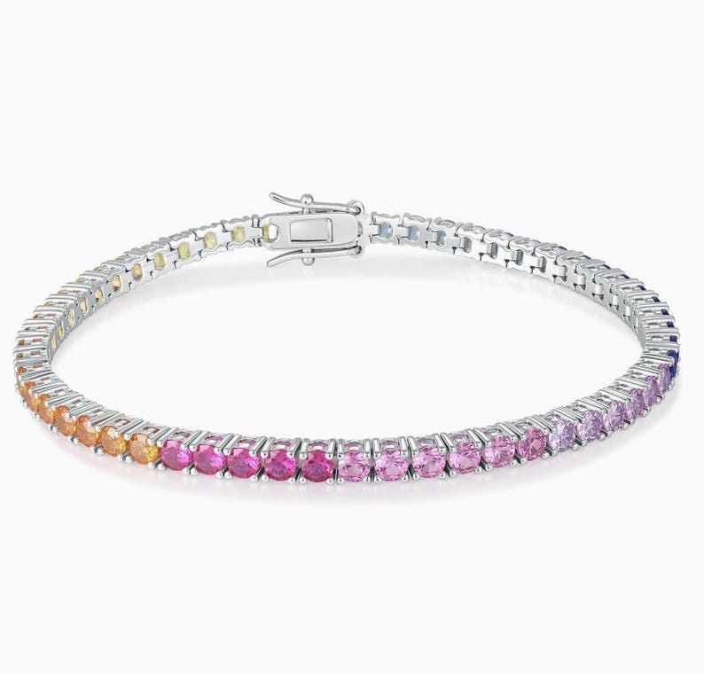May include: A silver-toned tennis bracelet featuring a continuous row of round-cut gemstones in a vibrant gradient color scheme, transitioning from orange and red to various shades of pink and purple, secured with a metal box clasp closure.