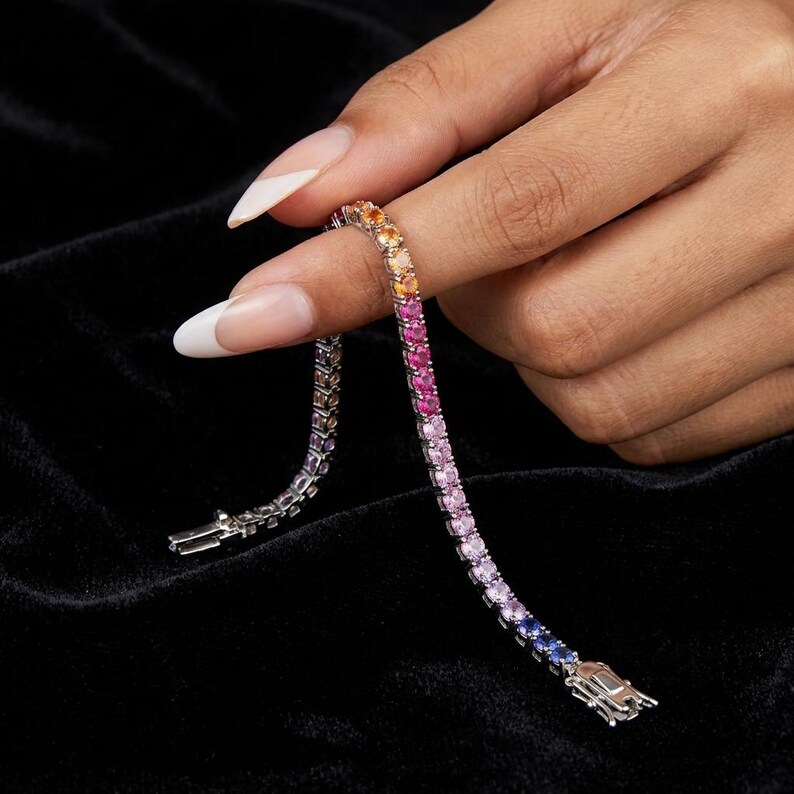 May include: A tennis bracelet featuring a gradient of round-cut gemstones in shades of orange, pink, purple, and blue, set in a metallic band. The bracelet displays a secure clasp mechanism at the end and a flexible, articulated link design.