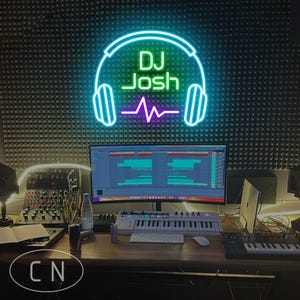 以下が含まれることがあります： 緑色の「DJ Josh」という文字がヘッドフォンの輪郭で囲まれたネオンサイン。その下に紫色の音波があります。サインはテクスチャ加工された壁に取り付けられています。木製の机の上には、コンピューターモニターと音楽機材があります。