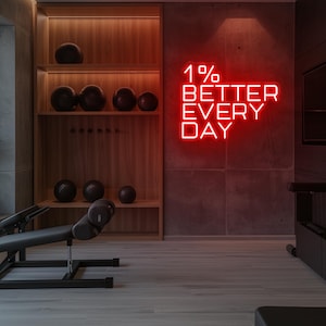 May include: A modern gym interior featuring wooden shelves with exercise balls and workout equipment. A red neon sign on the wall reads "1% BETTER EVERY DAY". The room has a minimalist design with a focus on fitness and motivation.