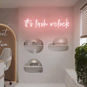 May include: A pink neon sign saying "It's lash o'clock" is fixed to a light grey wall. Below the sign are three white, organically shaped shelves displaying beauty products. A modern chair and a plant are also visible.