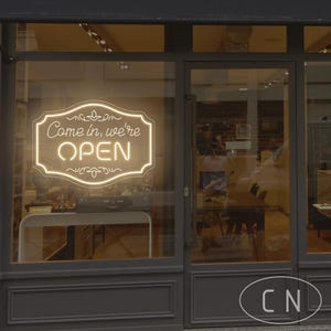 May include: A storefront with a neon sign in the window that reads "Come in, we're OPEN." The sign is in a decorative frame. The shop's exterior is painted gray, and the glass door reflects the interior.