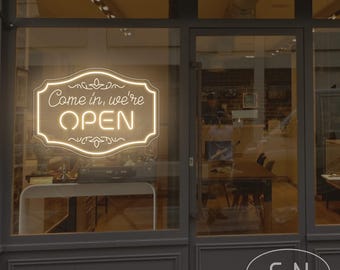 May include: A storefront with a neon sign in the window that reads "Come in, we're OPEN." The sign is in a decorative frame. The shop's exterior is painted gray, and the glass door reflects the interior.