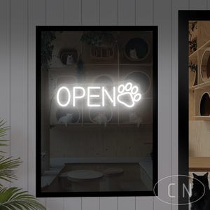 May include: A neon sign in a black frame displays the word "OPEN" with a paw print design. The sign is white and illuminates a cat-themed interior with white cats and wooden cat furniture.