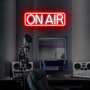 May include: A red neon "ON AIR" sign is mounted on a gray wall in a recording studio. The studio features a microphone, headphones, speakers, a computer monitor displaying audio editing software, and a keyboard.