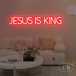 May include: A red neon sign that reads "JESUS IS KING" is mounted on a wall above a white sofa. The room is decorated with a round coffee table, flowers, and a floor lamp. The sign is the focal point.