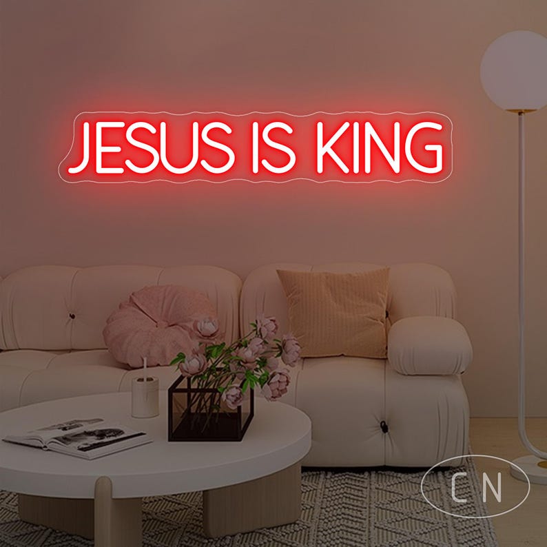 May include: A red neon sign that reads "JESUS IS KING" is mounted on a wall above a white sofa. The room is decorated with a round coffee table, flowers, and a floor lamp. The sign is the focal point.
