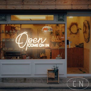 May include: Exterior shot of a shop with a glowing neon sign that reads "Open COME ON IN". The shop has a wooden facade and a glass window displaying various items. A small wooden sign is in front of the shop.