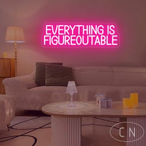 Puede incluir: Un letrero de neón rosa brillante con la frase "EVERYTHING IS FIGUREOUTABLE" ilumina una sala de estar. El letrero está montado en una pared sobre un sofá beige con cojines estampados. Una mesa de centro y una lámpara están en primer plano.