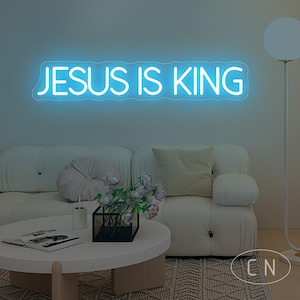 May include: A blue neon sign displaying the text "JESUS IS KING" is mounted on a wall above a white sofa. A round coffee table with a vase of flowers and a book is in front of the sofa. A floor lamp is on the right.