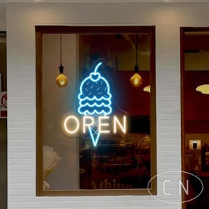 May include: A neon sign in a shop window features a blue ice cream cone with a cherry on top and the word "OPEN" in white. The sign is surrounded by a brown frame and hanging light bulbs.