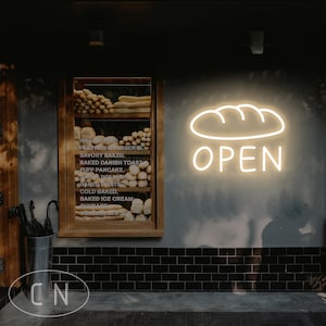 May include: Exterior shot of a bakery with a glowing neon sign that reads "OPEN" above a bread loaf outline. A wooden display case shows various baked goods. A metal bucket holds umbrellas. The wall is painted gray with black tile.