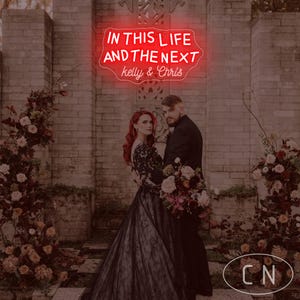 Może przedstawiać: Para pozuje przed ceglaną ścianą, otoczoną aranżacjami kwiatowymi. Neonowy znak nad nimi głosi "IN THIS LIFE AND THE NEXT" z nazwiskami "kelly & Chris" poniżej. Kobieta ma na sobie czarną suknię, a mężczyzna czarny garnitur.