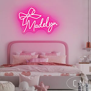 May include: A bright pink neon sign with the name "Madelyn" in a cursive font, topped with a bow design. The sign is mounted on a pink wall above a bed with pink bedding and pillows. The room has a soft, feminine aesthetic.