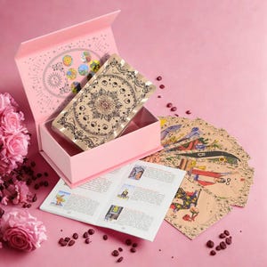 Puede incluir: Una caja rosa de cartas del tarot con una baraja de cartas con un diseño floral. La caja está abierta, revelando las cartas en su interior. Un folleto y cartas dispersas también son visibles sobre una superficie rosa.