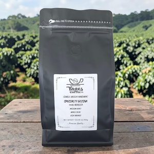 Puede incluir: Una bolsa de café negra con una etiqueta blanca que muestra "Yanes Coffee Co." y "Specialty Geisha". La bolsa está etiquetada como "Single Origin Honduras", "100% Arabica" y "USDA Organic". La bolsa está sobre una superficie de madera con un fondo verde.