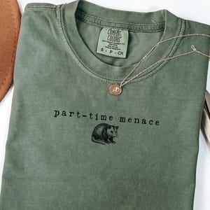 Pode incluir: Camiseta verde musgo com a frase "part-time menace" e um gráfico de um gambá. A camiseta tem gola redonda e mangas curtas. Um colar de ouro com um pingente está na camiseta.