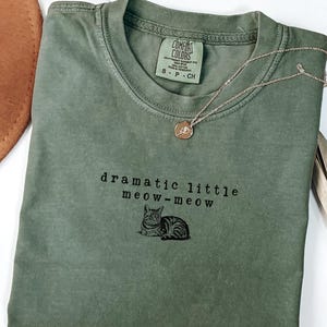 May include: A moss green t-shirt with the text "dramatic little meow-meow" and a cat illustration. A gold necklace with a pendant is draped on the shirt. The shirt is folded and displayed on a white surface.
