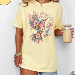 May include: A butter yellow short-sleeve t-shirt featuring a central graphic of a hummingbird surrounded by colorful floral and vine patterns. The shirt has a crew neckline and a relaxed fit. A circular badge in the top left corner says Butter.