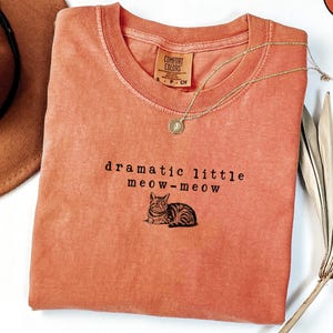 May include: A coral-colored t-shirt with the text "dramatic little meow-meow" and a cat illustration. A gold necklace and a brown hat are also in the image. The shirt is made of Comfort Colors material.