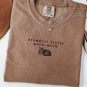 May include: An espresso-coloured t-shirt featuring the embroidered text "dramatic little meow-meow" above a small illustration of a tabby cat. The shirt has a crew neckline and a Comfort Colors brand label inside the collar.