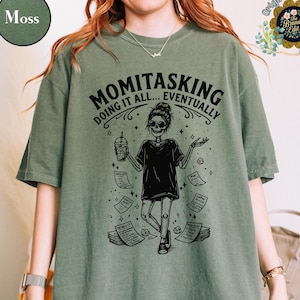 Könnte beinhalten: Moosgrünes T-Shirt mit dem Text "MOMITASKING DOING IT ALL... EVENTUALLY". Die Grafik zeigt eine Skelettfigur mit einer Kaffeetasse und Papieren. Das Shirt ist ein lässiger Stil für den Alltag.