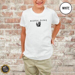 Puede incluir: Una camiseta blanca de manga corta con un gr&aacute;fico negro de un peque&ntilde;o mapache y el texto "mildly feral" impreso en una fuente estilo m&aacute;quina de escribir centrado en el pecho. La camiseta se combina con pantalones de color beige claro.