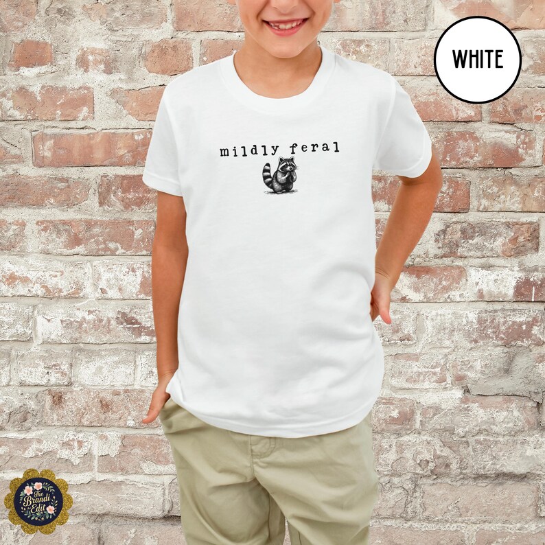 Puede incluir: Una camiseta blanca de manga corta con un gr&aacute;fico negro de un peque&ntilde;o mapache y el texto "mildly feral" impreso en una fuente estilo m&aacute;quina de escribir centrado en el pecho. La camiseta se combina con pantalones de color beige claro.