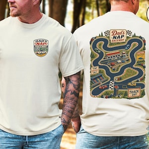 May include: Ivory-colored t-shirt featuring a colorful graphic of a race track with the text "Dad's Nap Raceway." The front of the shirt has a smaller version of the graphic. The back of the shirt has a detailed illustration of a race track with cars and buildings.