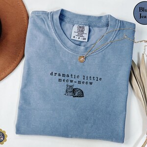 May include: A blue jean coloured t-shirt with a crew neckline and a Comfort Colors brand tag. The shirt features black embroidered text that reads dramatic little meow-meow above a small, detailed illustration of a tabby cat lying down.