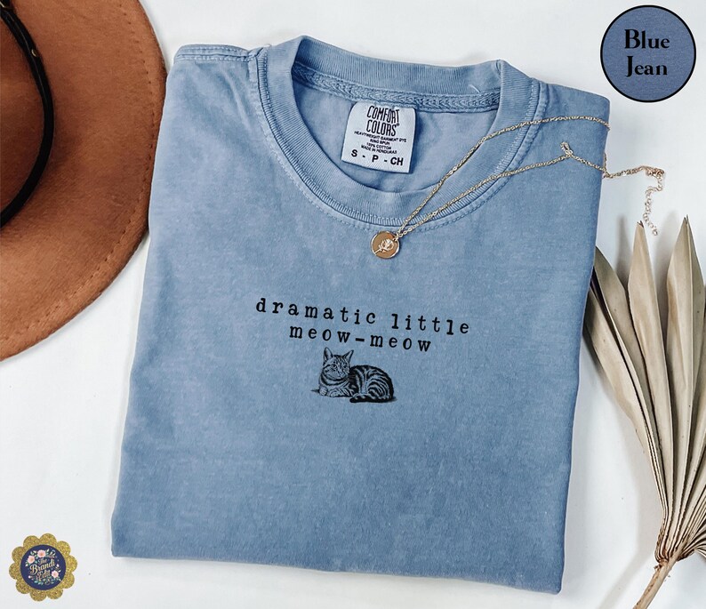 May include: A blue jean coloured t-shirt with a crew neckline and a Comfort Colors brand tag. The shirt features black embroidered text that reads dramatic little meow-meow above a small, detailed illustration of a tabby cat lying down.
