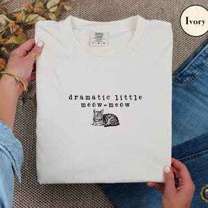 May include: An ivory t-shirt featuring a black graphic of a cat and the text "dramatic little meow-meow" printed in a typewriter-style font. The shirt has a crew neckline and a Comfort Colors brand label visible at the inner collar area.