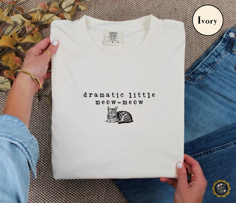 May include: An ivory t-shirt featuring a black graphic of a cat and the text "dramatic little meow-meow" printed in a typewriter-style font. The shirt has a crew neckline and a Comfort Colors brand label visible at the inner collar area.