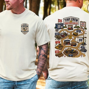 May include: An ivory t-shirt featuring a small logo on the front chest and a large graphic on the back. The logo reads Tiny Crew Construction Co. The back graphic depicts a construction site map with various vehicles, buildings, and signs.