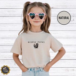 Puede incluir: Una camiseta beige de manga corta con un peque&ntilde;o gr&aacute;fico negro de un mapache y el texto "mildly feral" impreso en una fuente estilo m&aacute;quina de escribir en el centro del pecho. La camiseta tiene cuello redondo y un corte est&aacute;ndar.
