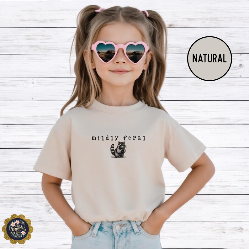 Puede incluir: Una camiseta beige de manga corta con un peque&ntilde;o gr&aacute;fico negro de un mapache y el texto "mildly feral" impreso en una fuente estilo m&aacute;quina de escribir en el centro del pecho. La camiseta tiene cuello redondo y un corte est&aacute;ndar.