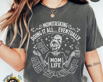 Może przedstawiać: Ciemnoszary t-shirt z białą grafiką szkieletu w opasce i okularach. Szkielet trzyma kubek z napisem Chaos Juice oraz listy zatytułowane Mom Life, Laundry i Pay Bills. Tekst głosi: Momitasking Doing It All... Eventually.